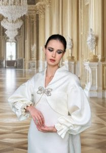 Una mujer que viste un sencillo vestido de novia con puños con volantes se encuentra en un salón adornado con detalles dorados, candelabros y pisos de madera pulida.