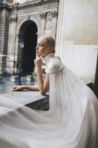 Una mujer con un vestido blanco de cuello alto su vestido de novia perfecto se apoya en una barandilla de piedra y mira pensativa a lo lejos, cerca de un edificio histórico.