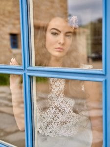 Una mujer con el pelo largo y un top de encaje, que recuerda la elegancia de un vestido de novia perfecto, está de pie detrás de una ventana azul, mirando al exterior con expresión neutra.