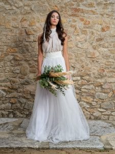 Una mujer con el vestido de novia perfecto, un traje de novia blanco sin mangas, está de pie contra un muro de piedra, sosteniendo un gran ramo de flores verdes y hierba de las pampas.