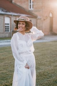 Una mujer con un vestido de novia perfecto un vestido blanco de encaje y un sombrero de ala ancha está de pie al aire libre sobre la hierba, sonriendo, con un edificio de ladrillo al fondo y la luz del sol brillando.
